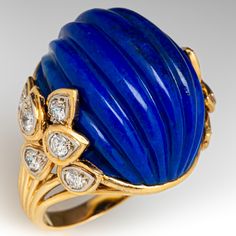 This exquisite cocktail ring is centered with one (1) oval shaped natural lapis lazuli with a carved fluted design. The shoulders of the ring are each accented with six (6) round brilliant cut diamonds bead set into petal shaped setting. The ring measures 23.6mm at the top, rises 15.8mm above the finger, tapering to 2.6mm wide and 1.2mm thick at the base of the shank. It is currently a size 7.25. Luxury Formal Blue Turquoise Ring, Luxury Lapis Lazuli Engagement Rings, Luxury Classic Domed Sapphire Ring, Luxury Gold Sapphire Fusion Ring, Elegant Blue Dome Ring For Anniversary, Elegant Blue Cabochon Dome Ring, Elegant Blue Gemstone Dome Ring, Luxury Lapis Lazuli Rings, Elegant Lapis Lazuli Ring With Polished Finish