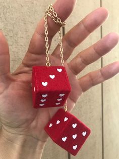 a hand holding two red dices with white hearts on them, hanging from a gold chain