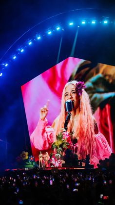 lady in pink outfit on stage with microphone and large screen behind her at night time