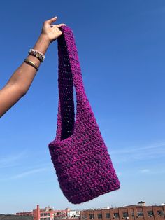 a hand holding up a purple crocheted bag in the air with buildings in the background