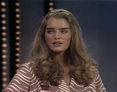 a woman with long hair sitting down in front of a tv screen and looking at the camera