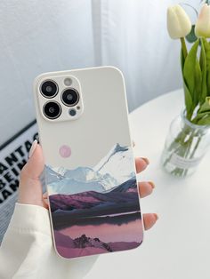 a woman is holding up her phone case with mountains on it and flowers in the background