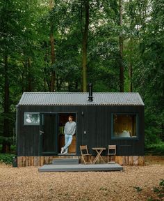 a man standing in the doorway of a tiny cabin surrounded by trees and grass with chairs around it