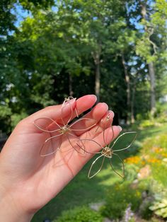 a hand holding two small metal spider webs in front of some trees and flowers