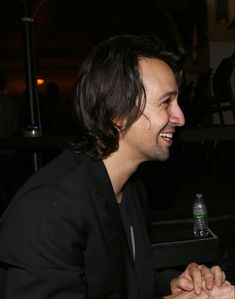 a man sitting at a table laughing and looking to the side with his hands clasped