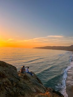 two people sitting on the edge of a cliff looking out at the ocean and sunset