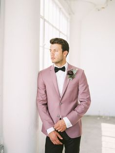 a man in a pink suit and bow tie standing next to a white wall with his hands in his pockets