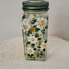 a glass jar with white flowers and green leaves painted on the lid is sitting on a lace tablecloth