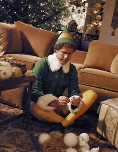 a man sitting on the floor in front of a christmas tree wearing an elf costume