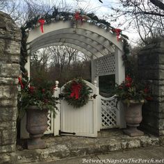 a white arbor with potted plants and wreaths on the top, in front of a stone wall