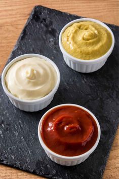 three small bowls filled with sauces on top of a black slate platter next to each other