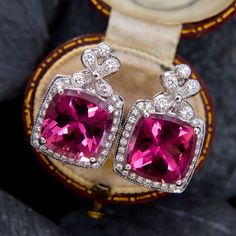 These collectable earrings are each accented with one (1) cushion cut natural Rubellite tourmaline set into a four-prong setting. The earrings are also each accented with thirty-two (32) bezel/ bead set round brilliant cut diamonds. The diamonds are bordered with milgrain edging. The earrings measure 19.5mm by 12.7mm and are finished with posts and friction backs for pierced ears. Elegant Cushion Cut Gemstones With Prong Setting, Luxury Tourmaline Jewelry With Prong Setting, Luxury Tourmaline Jewelry With Halo Setting, Classic Pink Gemstones For Formal Occasions, Elegant Tourmaline Jewelry With Halo Setting, Luxury Tourmaline Jewelry For Formal Occasions, Elegant Tourmaline Earrings With Gemstone Accents, Elegant Pink Gemstones With Accents, Classic Formal Earrings With Accent Stones