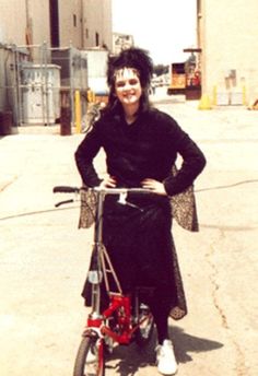 a woman standing next to a bike in the middle of a parking lot with buildings behind her