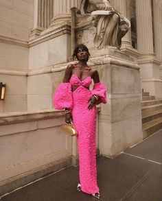 a woman standing in front of a statue wearing a bright pink dress and gold purse