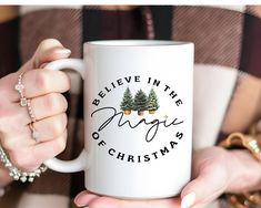 a woman holding a white coffee mug with christmas trees on it and the words believe in the magic of christmas