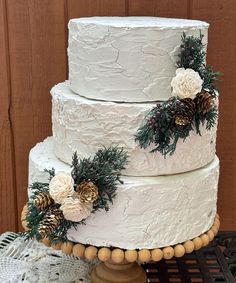 a three tiered white cake with pine cones and flowers on the top is sitting on a table