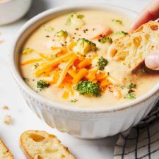 a person is dipping cheese into a bowl of broccoli and cheddar soup