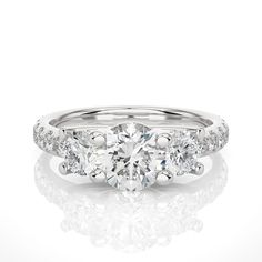 "This stunning brilliant trellis three stone engagement ring is a masterpiece of fine craftsmanship and exquisite design, perfectly combining the beauty of a 1.20+ carat Round lab-grown diamond with the sparkle of three stone diamonds. The Round diamond is of exceptional quality, with brilliant cut facets that maximize its natural radiance and fire. The diamond is set in a three stone shank design, which adds depth and dimension to the ring by framing the center stones appear to float invisibly above the surface of the ring. The ring's shank is also set with pave diamonds, which create a glittering band of brilliance that draws the eye toward the center stone. Each diamond is expertly set in a shared prong setting, which ensures that each stone is securely held in place while allowing maxi Classic Platinum Three Stone Rings, Classic Platinum Three-stone Ring, Three Stone Platinum Diamond Ring, Three Stone Diamond Ring In 14k White Gold, 14k White Gold Three Stone Diamond Ring, Luxury Three Stone 14k White Gold Rings, Formal Three Stone Round Cut Rings, Platinum Three Stone Ring With Round Band, Three Stone Platinum Round Rings