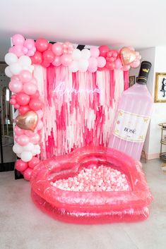 an inflatable heart shaped pool with balloons and streamers on the wall, next to a bottle of booze