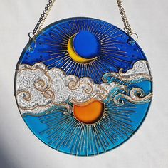 a glass sun and moon decoration hanging from a chain on a white wall with blue sky and clouds in the background