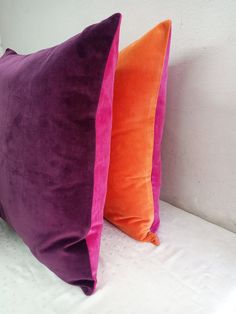 two purple and orange pillows sitting on top of a white bed next to each other