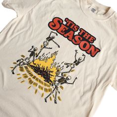 Get into the spirit of spookiness with our 'Tis The Season - Vintage Halloween T-Shirt! This one-of-a-kind, retro-inspired tee is perfect for those who love Halloween and autumn. The quirky graphic showcases a gang of skeleton pals boogying around a campfire. Our creative artists crafted this as a throwback to 1970's style, delivering a truly original shirt design. Indulge in unmatched comfort and style with our handcrafted t-shirts, featuring playful and unique designs. Proudly created in Penns 1970s Graphic Tee, Vintage Graphic Print T-shirt For Fall, Halloween Band Merch T-shirt With Graphic Design, Vintage Fall Graphic T-shirt, Band Merch T-shirt With Screen Print For Fall, Vintage Graphic T-shirt For Fall, Vintage Graphic Design T-shirt For Fall, Spooky Cotton T-shirt For Fan Merchandise, Fall Fan Apparel T-shirt With Graphic Print