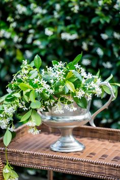 a vase filled with white flowers sitting on top of a wicker table next to a bush