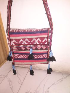 The bag features a blend of red, black, and beige colors, with intricate patterns and geometric shapes woven into the fabric. The use of goat and sheep wool gives the bag a durable yet soft texture, ideal for ladies' everyday use. The front flap of the bag is adorned with a series of small, black tassels, each accented with blue beads, adding a decorative touch. The strap is also intricately woven, matching the bag's overall color scheme and pattern. The craftsmanship suggests it may be of cultu Artisanal Design, Handcrafted Bags, Boho Bag, Boho Stil, Sheep Wool, Beige Color, Blue Beads, Soft Textures, Traditional Design