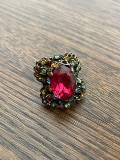 "Catch everyone's eye with this sparkling cocktail ring from the late 60s/70s.  A large pink glass stone flashes in the middle of the chunky ring, with a sprinkling of red, yellow, blue, green, and purple smaller baubles around it.  The scalloped design to the silver/pewter toned ring makes it almost look like a butterfly.  Adjustable band so you can wear it on any finger! Ring wiggles a little but is firmly attached.  Several of the small stones are missing, but the detailed design means it doe Vintage Multicolor Rings For Party, Vintage Jeweled Rings For Party, Vintage Red Crystal Gemstone Ring, Pink Jeweled Rings For Party, Pink Jeweled Party Rings, Retro Red Ring Jewelry, Sparkling Cocktail, Scalloped Design, Beige Hat