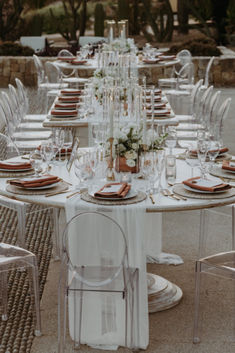 a long table set up with white linens and place settings for an outdoor dinner