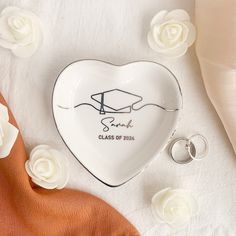 a white heart shaped plate with a graduation cap on it and two wedding rings laying next to it