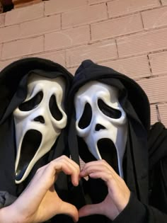 two people in black hoodies with white masks on their faces and hands making the shape of a heart