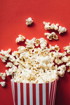 a red and white striped bucket filled with popcorn on a red background, surrounded by scattered popcorn kernels