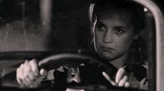 a woman driving a car with her hand on the steering wheel and looking at the camera