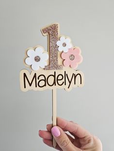 a hand holding a wooden cake topper with flowers on it that says madelyn