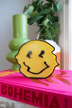 a yellow smiley face sitting on top of a pink book next to a green plant