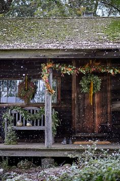 a wooden cabin with wreaths and decorations on the front porch during a snow storm