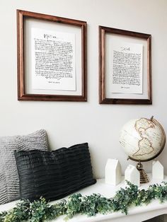 two framed pictures hang on the wall above a mantle with a globe and greenery