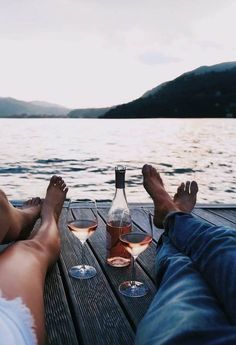 two people sitting on a dock with their feet propped up next to wine glasses in front of them