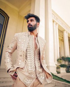 a man standing in front of a building wearing a white jacket and tan pants with gold sequins on it