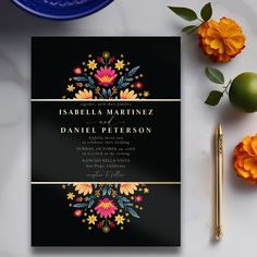 a black and gold wedding card with flowers on it next to a pen, orange flowers and a blue plate