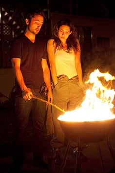 a man and woman standing next to a fire pit with flames in the dark behind them