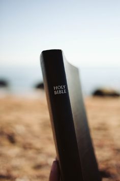 a hand holding up a bible in front of the camera on top of a beach