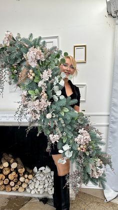 a woman holding a wreath in front of a fire place with logs and flowers around her