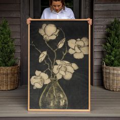 a woman holding up a painting with white flowers in a vase on the front porch