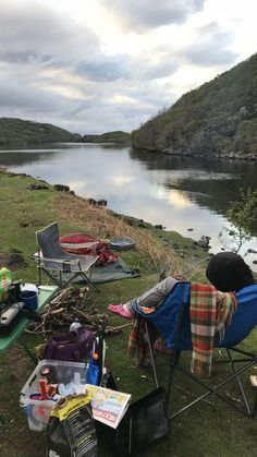 there is a lot of camping gear on the grass by the water and mountains in the background