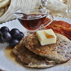 pancakes with syrup and bacon are on a white plate next to some grapes, bread and butter