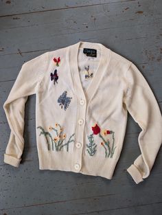 An adorable novelty sweater with blocky knit butterflies flitting over flowers and grasses. In very good condition. 15 inches across the shoulders, 16 inches across the bust. 20 inches long. White Embroidered Knit Cardigan, Flower Patch Cardigan, Long Sleeve Knit Cardigan With Floral Embroidery, White Vintage Cardigan With Floral Embroidery, Vintage Cream Embroidered Cardigan, Novelty Sweater, Grasses, Cardigans For Women, Sweaters & Cardigans