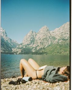 a woman laying on top of a beach next to a body of water with mountains in the background