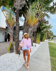 a woman in a pink shirt and blue shorts is standing on the sidewalk near palm trees